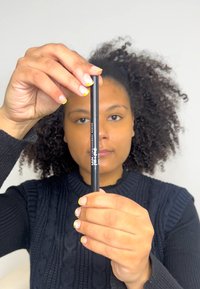 Svart eyelinerpenna hålls mellan fingrarna, med en slät yta och en smal design. Förpackningen inkluderar vit varumärkestext.