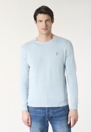 Uomo che indossa un maglione azzurro chiaro a maniche lunghe con un piccolo logo sul petto e jeans blu, in piedi davanti a uno sfondo chiaro e uniforme.