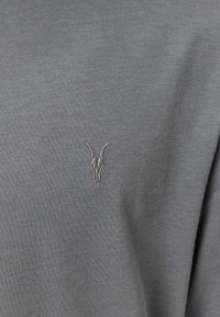 Szary bawełnowy t-shirt z delikatnym, haftowanym wzorem czaszki z rogami w jaśniejszym odcieniu szarości po lewej stronie klatki piersiowej.