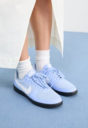 Ben iført hvide sokker og lyseblå Nike sneakers med hvid swoosh, stående på et blåt tæppe ved siden af et hvidt stof.