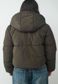 Puffermantel in dunkelolivgrün. Mit Kapuze, gestepptem Design und kurzem Schnitt. Kombiniert mit hoch taillierten blauen Jeans.