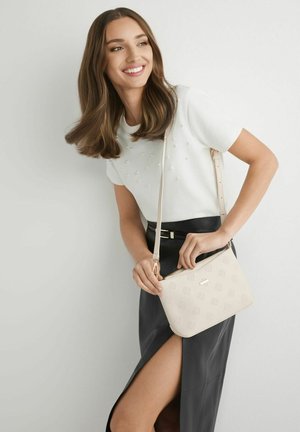 Kobieta o brązowych włosach do ramion uśmiecha się, ubrana w biały top z perełkami, czarną spódnicę z rozcięciem na uda, trzyma beżową torebkę na ramię.