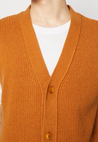 Rippweste in Orange mit V-Ausschnitt, ausgestattet mit drei Holzknöpfen und einem weißen Rundhalsausschnitt darunter.