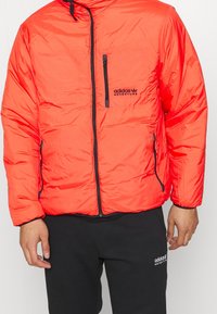 Orange puffjacka med en dragkedjeficka och svarta detaljer. Har en hög krage och en lätt, quiltad design.