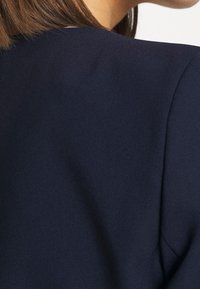 Dark navy fabric details with a smooth texture, featuring a clean silhouette and structured shape at the shoulders. No additional accents visible.