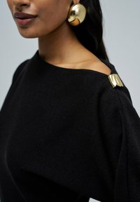 Suéter negro con un escote amplio. Presenta un acento dorado en el hombro y una textura suave. Grandes pendientes redondos dorados visibles.