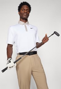 Polo branco de mangas curtas com colarinho bege, logotipo da Nike em preto. Calças bege, cinto preto, segurando um taco de golfe com uma luva branca.