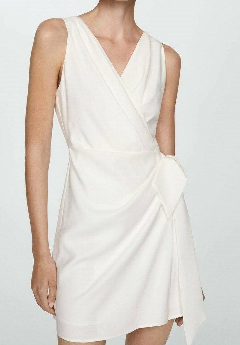 Robe portefeuille blanche sans manches avec un décolleté en V et un nœud sur le côté, portée par une personne sur un fond clair uni.