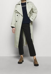 Trench coat grigio chiaro con bottoni neri, felpa blu navy con logo, pantaloni neri cropped e scarpe stringate marroni. Texture lisce.