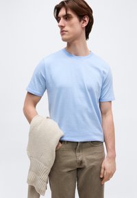 Νέος άνδρας με ανοιχτό μπλε κοντομάνικο πουκάμισο και καφέ παντελόνι, κρατώντας ένα μπεζ πλεκτό πουλόβερ στο αριστερό του χέρι.