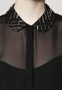 Blusa nera in tessuto trasparente, con colletto geometrico in perline nere e metalliche e chiusura con bottoni davanti.