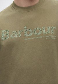 Μπλουζάκι χακί-πράσινο με τη λέξη "Barbour" με μεγάλα διακοσμητικά γράμματα και τη φράση "Κατασκευαστές ρούχων για υπαίθριες δραστηριότητες" σε μικρότερη γραμματοσειρά από κάτω.
