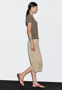 Donna che cammina in una gonna midi beige, top a maniche corte verde oliva, sandali rossi e bracciale in argento su uno sfondo bianco semplice.