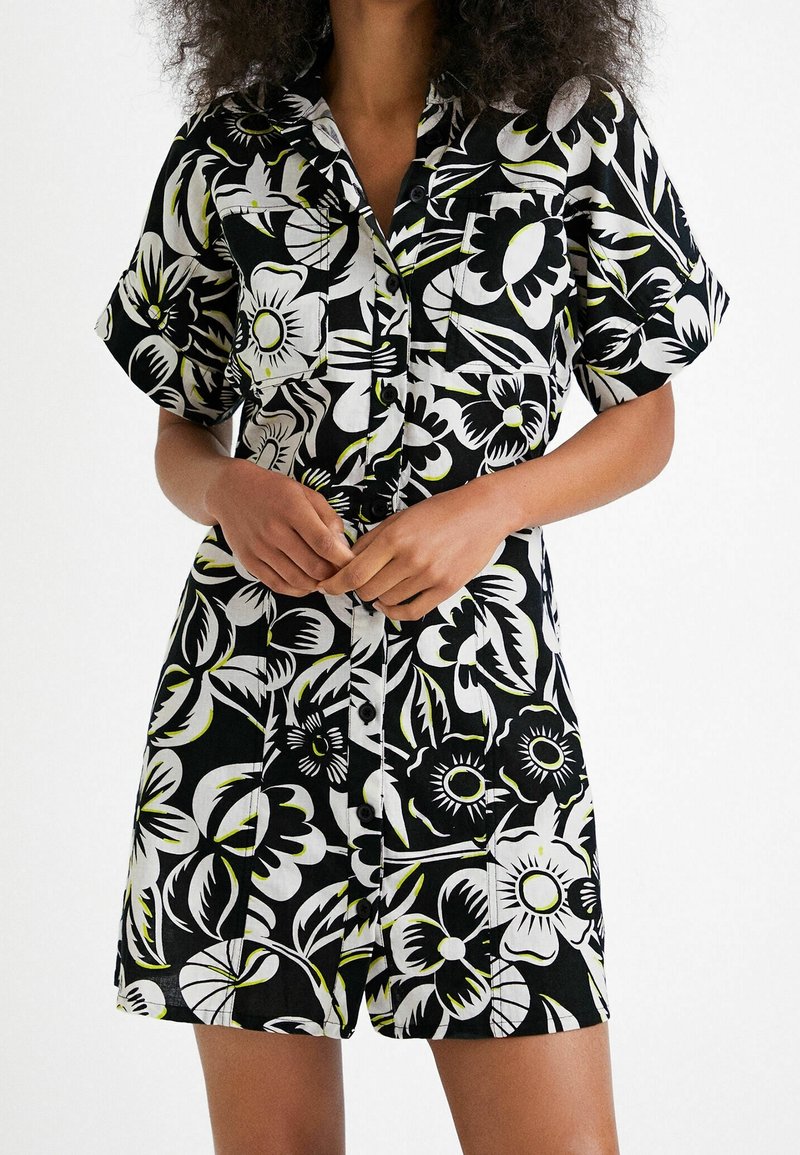Femme portant une combinaison short à manches courtes à boutons, noire et blanche à motif floral, avec des poches poitrine avant, debout devant un fond blanc uni.
