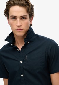 Camisa de manga corta, con botones, en azul marino oscuro con un pequeño bolsillo en el pecho. Presenta un cuello alzado y botones blancos. Textura de tela suave.