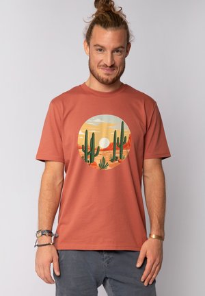 Roodbruin katoen t-shirt met een circulaire woestijnscène afdruk van cactussen en een zon, tegen een gedempte luchtachtergrond.