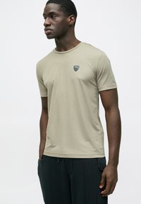 Kaki rövid ujjú póló kerek nyakkal és kis fekete logóval a mellrészen, sima, könnyű anyagból készült.