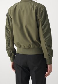 Olivově zelená bomber bunda s matným povrchem, s vycpanými saténovými rukávy, žebrovanými manžetami a pasem, zobrazena zezadu.
