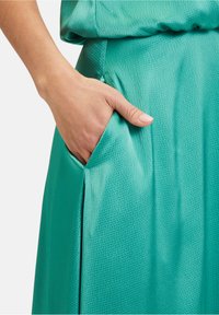 Turkusowa spódnica o fakturowanej powierzchni z boczną kieszenią i marszczonym pasem. Materiał ma subtelny połysk i wzorzystą powierzchnię.