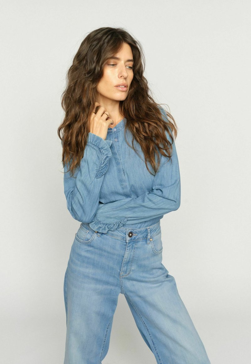 Mujer con cabello largo y ondulado, vestida con una blusa vaquera azul claro con puños fruncidos y jeans de talle alto, posando frente a un fondo claro y liso.