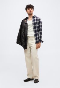 Νεαρός άνδρας με κρεμ παντελόνι, λευκή φανέλα, μισοφόρεμο καρό πουκάμισο και μαύρο δερμάτινο μπουφάν περασμένο πάνω από τον έναν ώμο, στέκεται μπροστά σε λευκό φόντο.