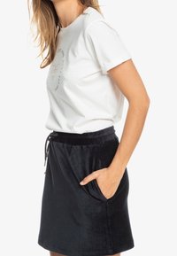 Weißes Baumwoll-T-Shirt mit hochgekrempelten Ärmeln und rundem Logo auf der Brust, kombiniert mit einem dunklen Velours-Minirock mit elastischem Taillenbund und Taschen.