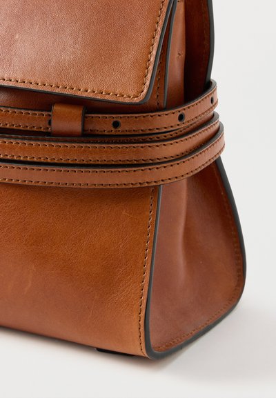 Sac à main en cuir marron avec détails cousus et deux fines lanières enroulées autour du devant sur un fond blanc.