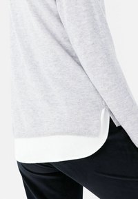 Hellgrauer Pullover mit strukturiertem Finish, mit seitlichen Schlitzen, über einem weißen Hemd getragen. Kombiniert mit dunklen gerippten Hosen.