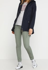 Marinblå vattentät jacka med huva, dragkedja och två framfickor. Matcher med smala olivgröna byxor och vita sneakers. Avslappnad outfit.