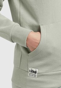 Mano inserita nella tasca anteriore di una felpa grigia chiara con polsini a coste e etichetta del marchio vicino all'orlo.