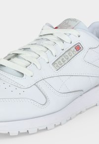 Λευκό δερμάτινο αθλητικό παπούτσι Reebok με λευκά κορδόνια, λογότυπο Reebok και μικρή βρετανική σημαία στο πλάι, εμφανιζόμενο σε κοντινή γωνιακή λήψη.
