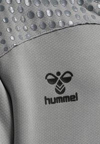 Grå atletisk tyg med en texturerad övre del som har glansiga runda prickar. I mitten finns en svart logotyp som säger "hummel."