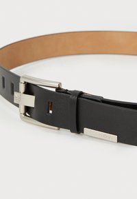 Ceinture en cuir noir avec une texture lisse, bouclerie en métal argenté et un détail rectangulaire au logo Calvin Klein. Possède des trous ajustables.