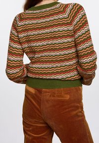 Stickad tröja med ett zigzag-mönster i jordnära toner av grönt, orange och vitt. Ribbad grön nederkant och ärmar, i kombination med bruna manchesterbyxor.