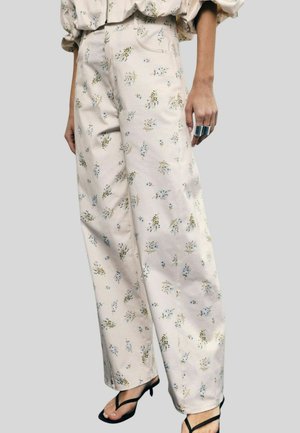 Donna che indossa pantaloni crema a vita alta e gamba larga con una piccola stampa floreale blu e verde, abbinati a sandali neri e un anello d'argento sulla mano destra.