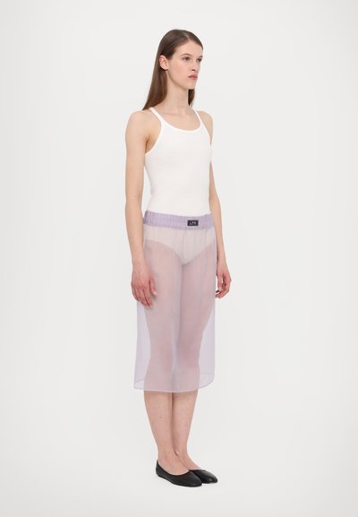 Jupe lavande transparente avec une taille élastique et un logo noir. Portée par-dessus un débardeur blanc, elle a une texture fluide et douce.