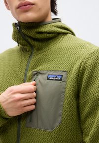 Άτομο που φοράει πράσινο φλις μπουφάν Patagonia με υφή και φερμουάρ σε τσέπη στο στήθος, το οποίο ανοίγεται με το χέρι.