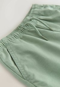 Pantaloncini verde chiaro realizzati in tessuto liscio con cintura elastica, coulisse e tasche laterali. Sono visibili dettagli cuciti.
