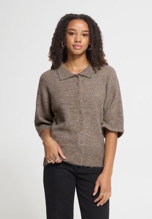 Femme aux cheveux bouclés portant un cardigan marron boutonné à manches courtes et un pantalon noir, debout devant un fond uni clair.