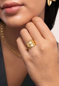 Goldblatt-förmiger Ring an einer Hand, mit strukturierten Details und einem leicht offenen Design. Glatte Hauttöne und dezenter Schmuck ergänzen den Look.