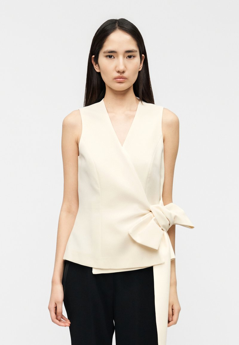 Ασύμμετρο κρεμ τοπ τύπου wrap από δομημένο ύφασμα, με μεγάλο φιόγκο στη μέση και βαθύ V λαιμόκομμα. Σετ με μαύρο παντελόνι.