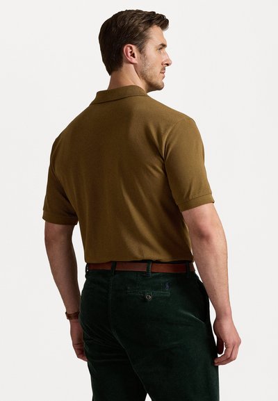 Camisa polo castanha de manga curta, em tecido de malha texturizada, usada com calças de veludo cotelê verde-escuro e um cinto de couro castanho.