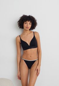Svart trådløs bh med dyp V-hals og justerbare stropper, kombinert med matchende svart bikinitruse med hvit midjebånd.