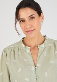 Blusa verde claro con un escote fruncido, botones en el frente y bordado de hojas blancas en toda la tela. Material suave y texturizado.