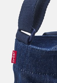 Denim bag in dark blue with textured surface, featuring a fabric strap and a red Levi's tag. Includes visible stitching and hardware details.