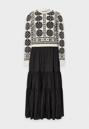 Une femme porte un pull couleur crème à motifs géométriques noirs, une longue jupe noire à volants, des mocassins noirs et un sac porté à l’épaule marron.