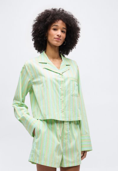 Γυναίκα με σγουρά μαλλιά, που φοράει ανοιχτό πράσινο ριγέ πουκάμισο με κουμπιά και αντίστοιχο σορτς, στέκεται με το ένα χέρι στην τσέπη.