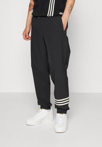 Czarne spodnie sportowe z tapered leg i elastycznymi mankietami, z trzema białymi paskami na dolnym obszyciu. Szczegół z zamka błyskawicznego w bocznej kieszeni.