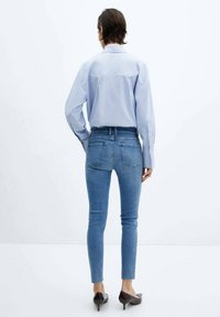 Camisa de botón azul claro de mangas largas, combinada con jeans ajustados azul y zapatos de tacón marrones, vista desde la espalda.