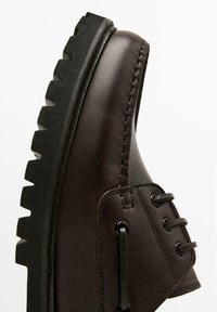 Scarpa in pelle marrone con suola nera spessa, caratterizzata da lacci rotondi e dettagli di cuciture laterali. La vista laterale mette in risalto i materiali strutturati.